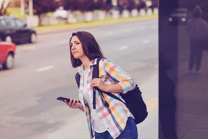 A woman with a cell phone waiting for a bus or car by the side of a street.