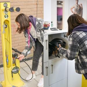 A collage of two images side by side. The image on the left shows a woman next to the yellow bike repair stand. The image on the right shows the same woman putting a helmet into a square locker.