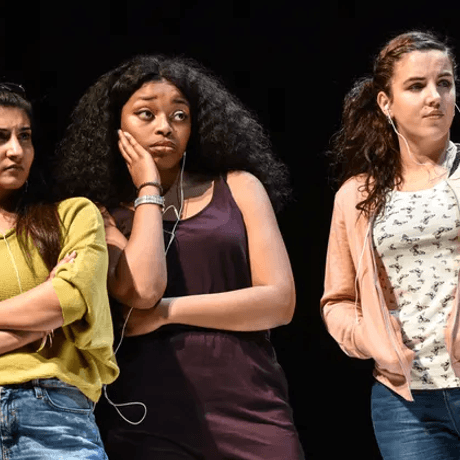 Three young women stand on stage looking judgementally to a character out of shot