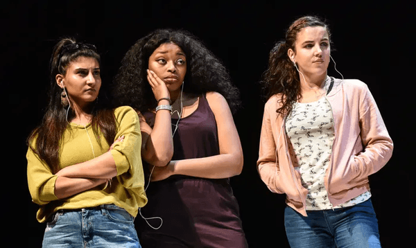 Three young women stand on stage looking judgementally to a character out of shot