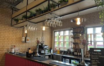 The bar area. There paper straws and wooden cutlery in a cup on the left. There are two beer taps behind the counter. There is a coffee machine and selection of teas behind the taps. Behind them is a window. There are wine glasses hanging above the counter, as well as some plants.
