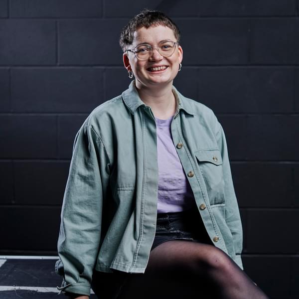 Portrait of Ali in front of a black brick wall. She is wearing a blue denim jacket over a purple t-shirt. She is also wearing a pair of glasses and silver hoop earrings.