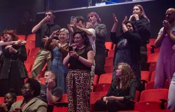 A group of audience members - some of them are standing and applauding, some remained seated and one is taking a photo of the stage on their phone,
