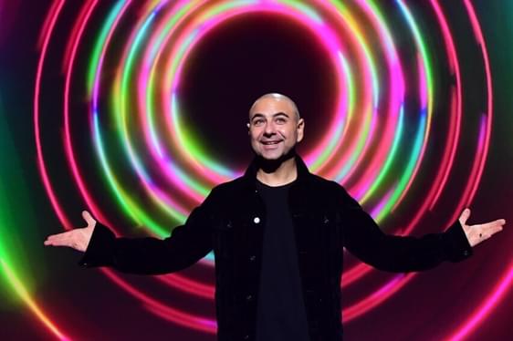 Joe Avati stood on stage with his arms open, in front of a backdrop of multi-coloured circles.