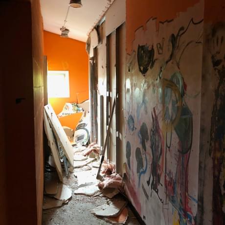 A hallway in reconstruction. The walls are orange and the wall on the right is in deconstruction. There are wooden planks, cardboard and pieces of the wall on the floor,