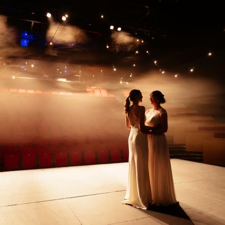 the stage and auditorium with red seats with a couple standing on the stage facing each other holding hands in white dresses. There is a vintage effect on the image