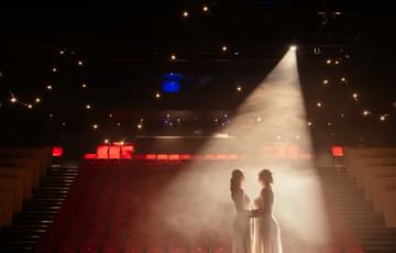 the stage and auditorium with red seats with a couple standing on the stage facing each other holding hands in white dresses with a bright spot light shining down on them