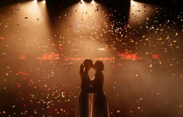 the silhouette of two women kissing on stage in white bridal dress. They are surrounded by confetti falling from above
