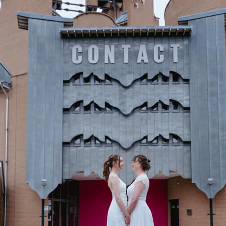 Two women stood in front of the Contact building, holding hands and look at each other in white bridal dresses