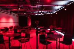 A space to hire set up with red lighting and lots of small tables with chairs around each and black curtains along the right hand side