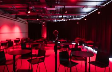 A space to hire set up with red lighting and lots of small tables with chairs around each and black curtains along the right hand side