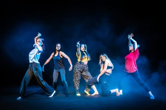 A group of dancers stand on a smokey blue lit stage