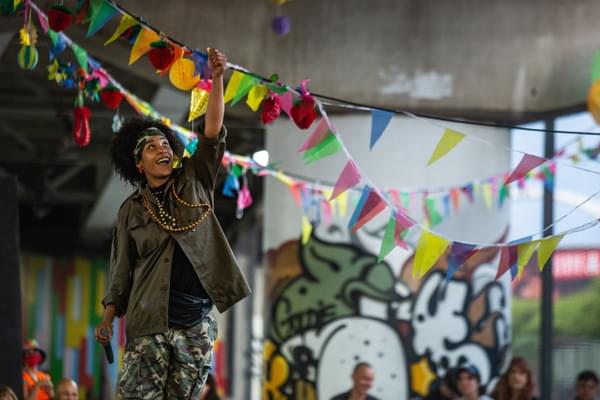 A young person stands in a skatepark adorned with graffiti and bunting, looking up and smiling at a lit lighter in their hand