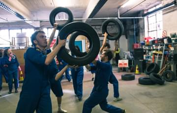 A group of people in a workshop, wearing blue overalls, carrying car tyres about their heads