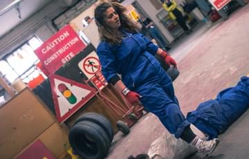A person wearing blue overalls is stepping over another person led face down on the floor, in a workshop setting