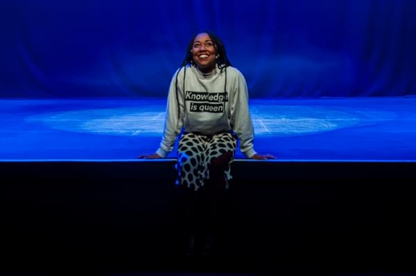 Keisha Thompson sat on the edge of a stage. The stage is lit blue. Her hair is long, braided and she is wearing a grey sweatshirt that says "Knowledge is queen".