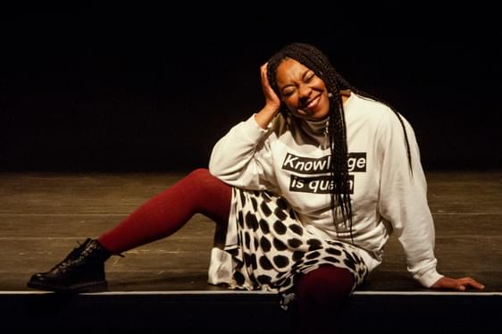 Keisha Thompson sat on the edge of a stage, she is wearing red tights, a polka dot skirt and a jumper that says knowledge is queen