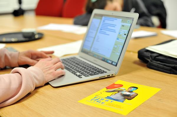 A pair of hands typing on a laptop. Next to the laptop is a yellow flyer for Dibby Theatre's First Time.