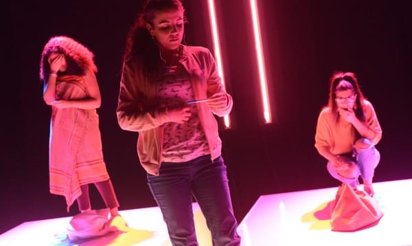 Three young woman perform on stage lit by neon pink lights