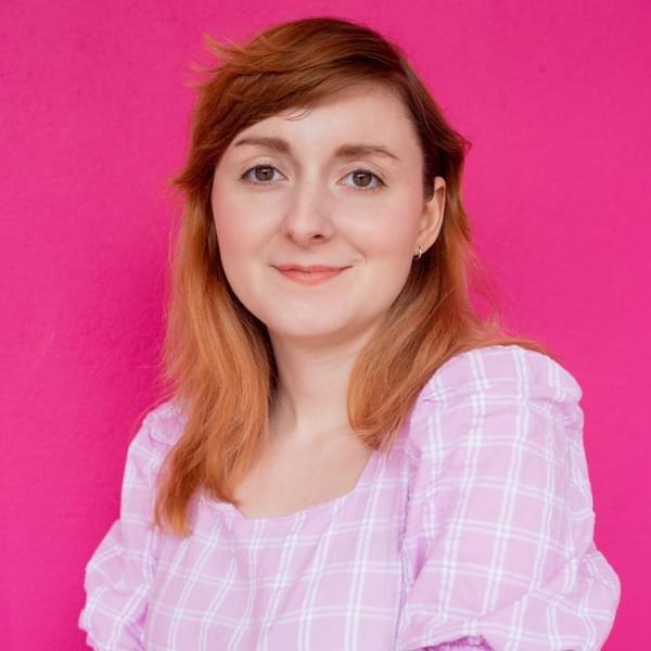 A young person with red hair wears a pink checked dress against a hot pink background