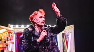 Luke Hereford stood in front of a lit up wardrobe. His hair is pink and dressed in a black fur coat. He is holding a mic in his right hand and holding up his left hand.