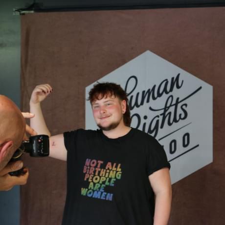 person in front of a sign which says human rights tattoo holding their arm up that has the letter tattooed on smiling at the person taking a photo