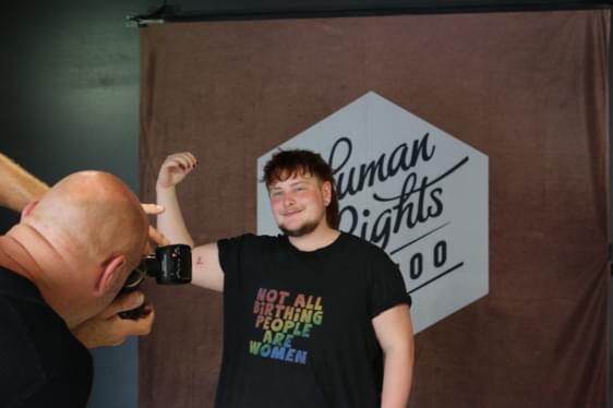 person in front of a sign which says human rights tattoo holding their arm up that has the letter tattooed on smiling at the person taking a photo