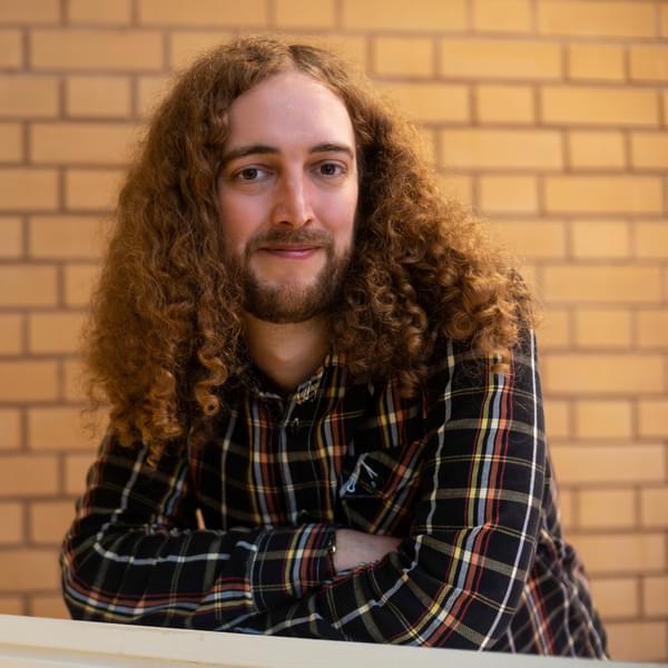 A young person with long curly hair and a checked shirt smiles wistfully at the camera