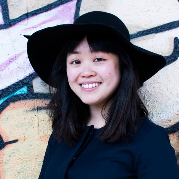 Headshot of Jo Yee Cheung in front of a graffitied wall. Her hair is shoulder length and black. She is wearing a black t-shirt, a black hat and smiling.