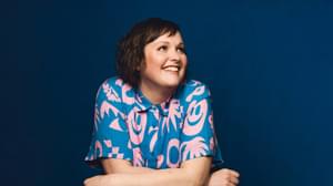 Josie Long is looking to her left and smiling, arms crossed. She is in front of a dark blue background and wearing a blue and pink shirt.