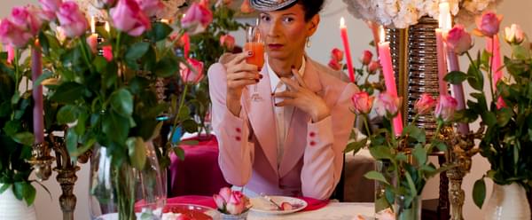 Nando Messias seated at a table holding a glass, wearing pink blazer and a bright fascinator. They are surrounded by flowers and candles.