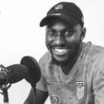 Black and white portrait of Paul Olubayo. He is wearing a cap and smiling with a recording microphone next to him.