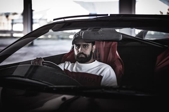 A man sits in the front seat of a car, driving and staring down the camera