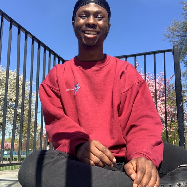Pelumi is sitting outdoors on the floor in front of a black fence. There are some trees behind him and the sky is bright and blue. He is wearing a red sweatshirt, black jeans, black boots and black silk scarf.