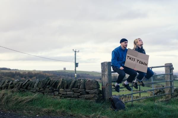 A man and woman sit on a fence in a field holding a cardboard sign reading THIS TOWN