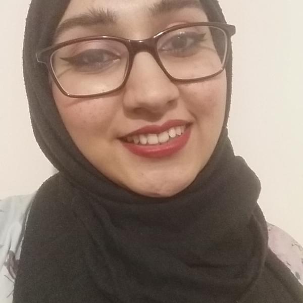 A smiling selfie of Sadia, they are wearing glasses, a black headscarf and red lipstick.