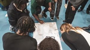 A group of young people huddle on the floor, drawing on a shared sheet of paper