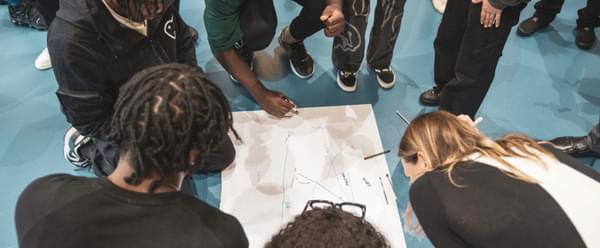 A group of young people huddle on the floor, drawing on a shared sheet of paper