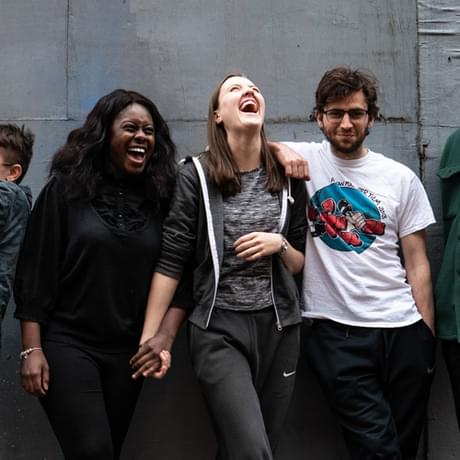 A diverse group of young people from Contact Young Company (CYC) stand against a wall laughing and smiling.