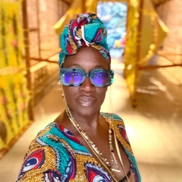 Headshot of Theresa, wearing a multi colored headscarf and dress, with necklaces and large blue sunglasses