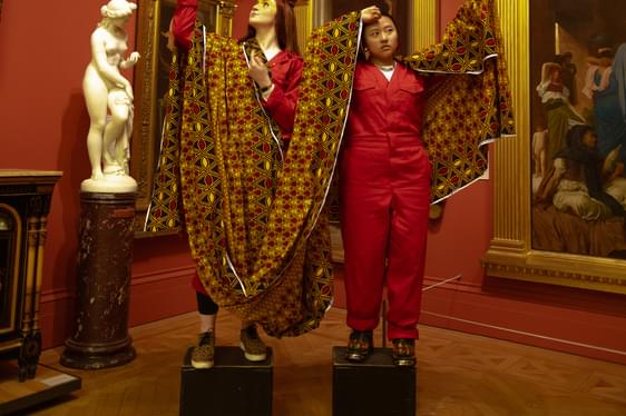 Two people stood on pedestals in an art gallery setting. They are wearing red overalls and draped in a golden sheet. There is tray with tea and biscuits on the floor in front of them.