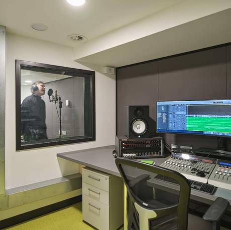 recording studio for music with person standing in the booth and the long screen computer in the room in front of it