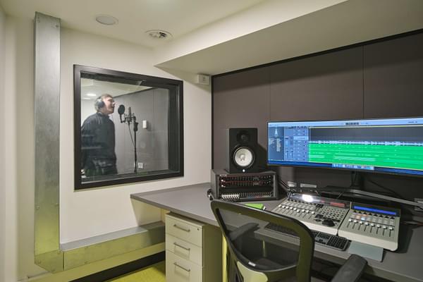 recording studio for music with person standing in the booth and the long screen computer in the room in front of it