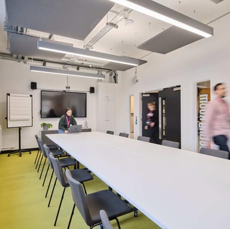meeting room with long conference table in the middle and large screen at the end of the room. people walking into the room as if they are about to start a meeting