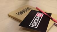 A Contact notebook, Contact pen and Contact pencil stacked on a wooden desk.