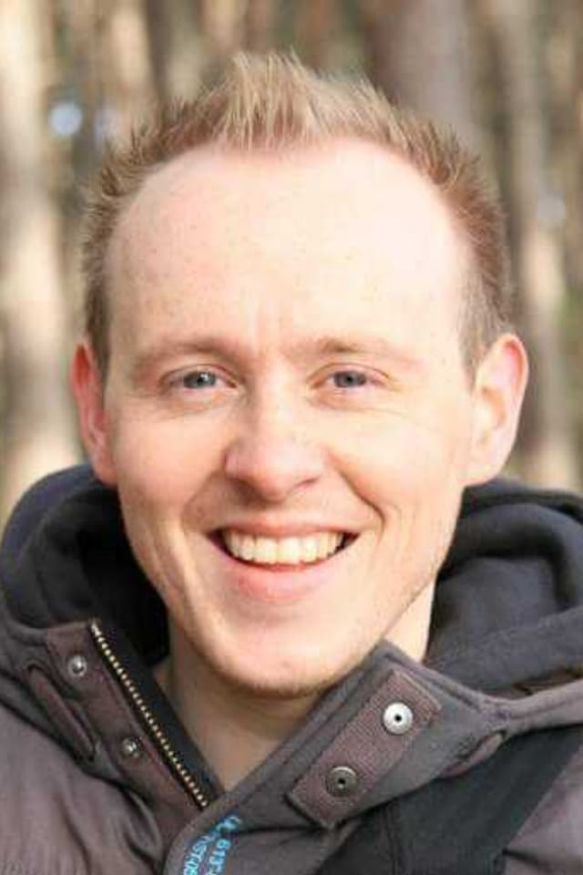 A smiling portrait of a young man in his coat in the woods.