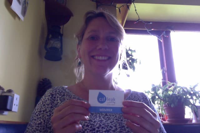 A woman in her home holding a CSF Leak Association Membership card.