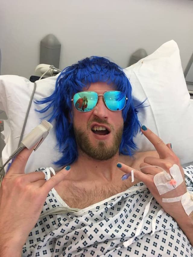 A man lying in bed with a blue wig, sunglasses, and making the rock-on hand symbol.
