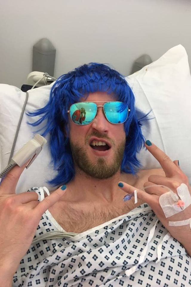 A man lying in bed with a blue wig, sunglasses, and making the rock-on hand symbol.