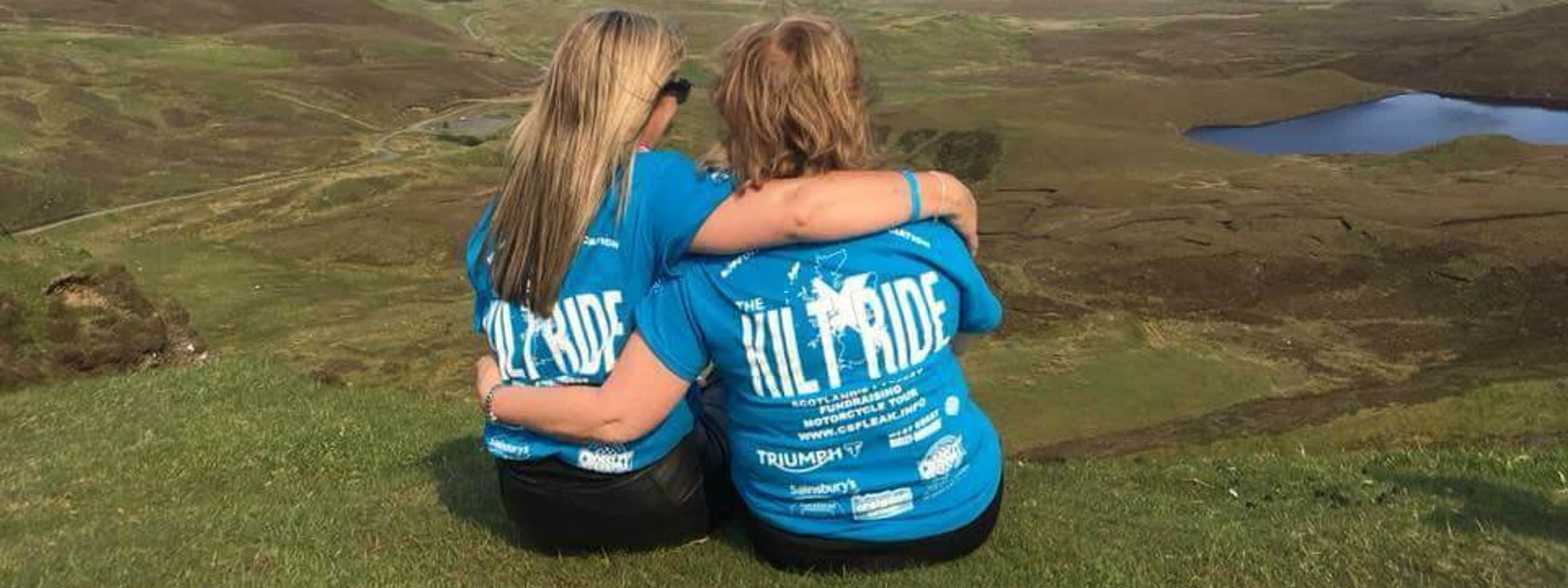 A photo of two females sitting and hugging each other sideways on a beautiful hill.